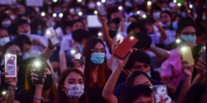 The image shows a large crowd of K-pop fandom culture in action, with dedicated fans holding up their smartphones with flashlights on, creating a sea of lights at what appears to be a concert or fan event. Fans, many wearing masks, are demonstrating K-pop fan power through their synchronized participation, illustrating the massive fandom impact on K-pop events through their collective light display, a common practice during ballads or emotional moments at K-pop concerts.