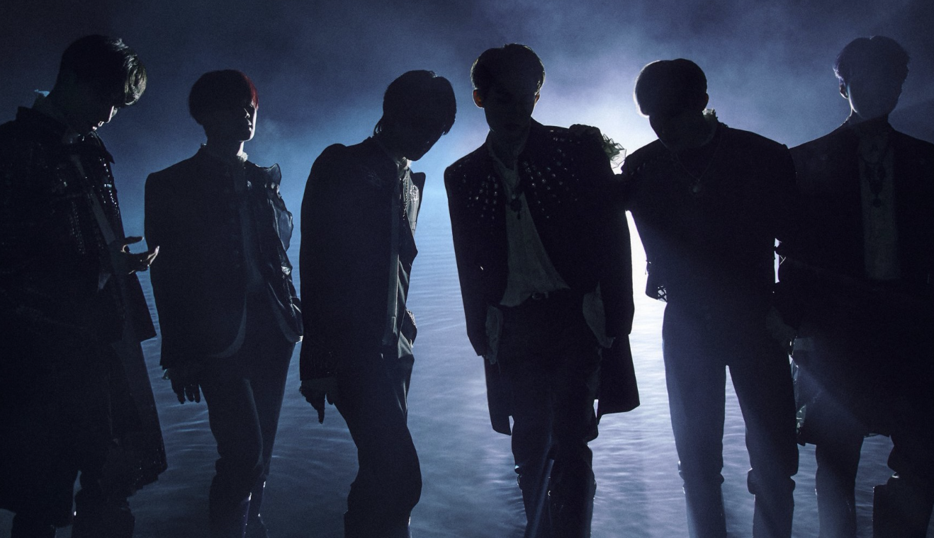Silhouettes of a top K-pop group standing in dramatic backlit pose against a blue-toned smoky background, highlighting the artistic visual direction that defines K-pop groups 2025 will showcase. The powerful staging and mysterious atmosphere represent the evolution of K-pop performance aesthetics, capturing the anticipation surrounding major groups' comebacks and artistic reinvention in the 2025 K-pop landscape.