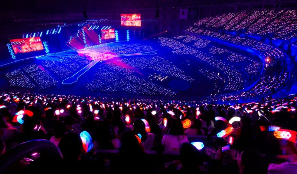 K-pop Idol System concert stadium filled with fans holding colorful lightsticks at a major idol group performance, showcasing the massive scale of K-pop fandom compared to Hollywood celebrity events