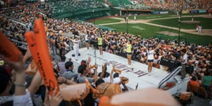 This image captures the vibrant atmosphere of a Korean baseball stadium, showing cheerleaders performing on a dedicated platform while enthusiastic fans participate in coordinated cheering. In the foreground, fans can be seen waving orange thunder sticks, a popular cheering item at KBO games. The cheerleaders, dressed in bright yellow tops and shorts, are leading the crowd in synchronized chants and movements, while the baseball game continues on the field in the background. The stands are filled with a diverse crowd, demonstrating the inclusive and festival-like nature of Korean baseball culture. This scene perfectly illustrates the unique fan experience that makes Korean baseball different from other leagues around the world, where entertainment and active participation are central to the stadium experience.