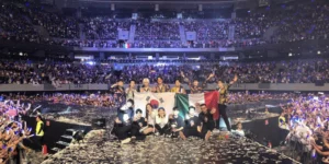 K-pop group performing at a packed stadium in Mexico, holding both Korean and Mexican flags on stage with confetti covering the floor