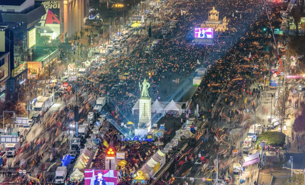 Aerial night view of massive K-protest culture demonstration in Seoul featuring thousands of peaceful protesters with light sticks creating a festival-like atmosphere around Gwanghwamun Square