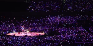 A Coldplay concert in South Korea showing thousands of colorful light sticks illuminating the dark stadium while the performer plays on a circular stage at the center