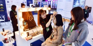 Professional makeup artist applying K-beauty cosmetics to visitor at K-Beauty Festival 2025 exhibition booth with Hollywood-style vanity mirror and cosmetic display